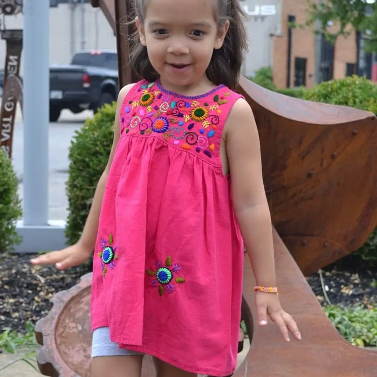 Young girl wearing a pink dress with colorful embroidery standing outdoors.