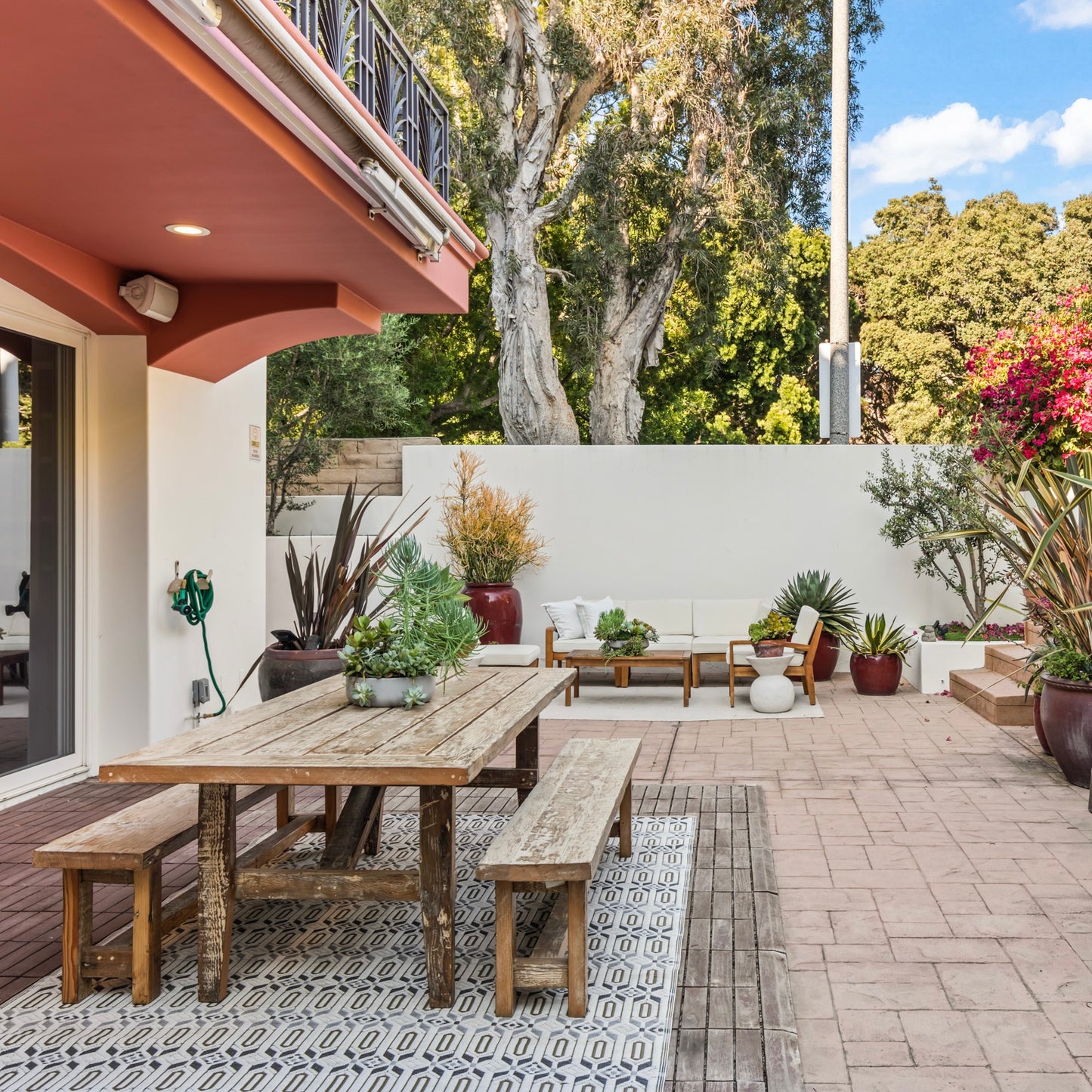 Outdoor patio area with wooden table, benches, and seating area with plants and a white wall.