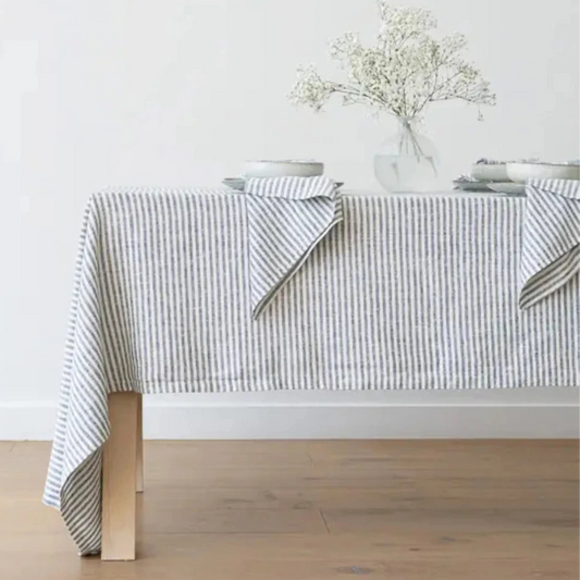A striped linen tablecloth in white and blue, draped over a table with wooden legs. The table is set with white plates and a vase with flowers.