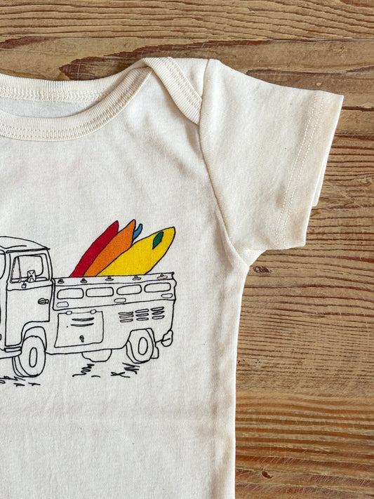 White baby onesie with a graphic of a truck carrying surfboards on a wooden surface