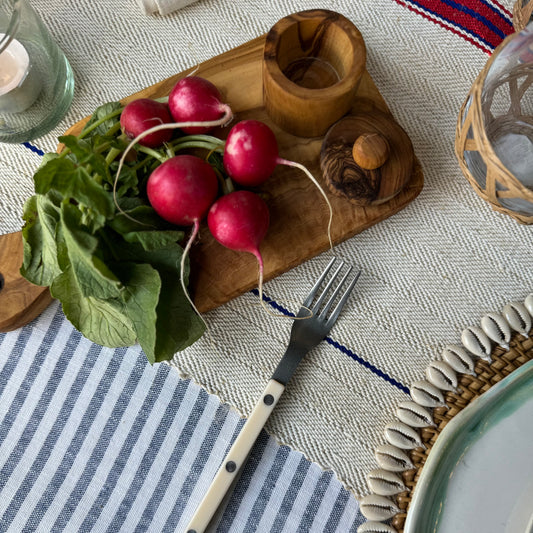 An olive wood paddle board used as a cutting board with radishes being cut on it, placed on a table with a striped blue and white tablecloth.