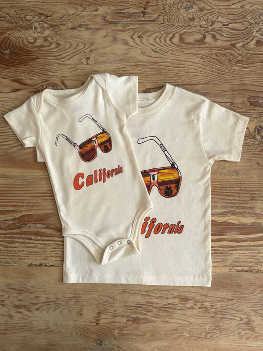 Two baby onesies with sunglasses and 'California' text on a wooden surface