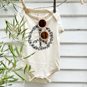 light pink toddler sunglasses hanging on a cream colored onesie with a floral peace sign on the front