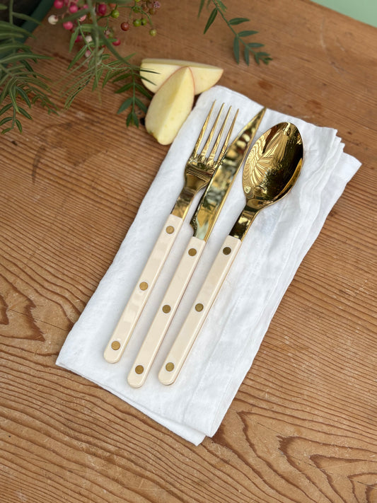 Gold-toned flatware set with cream-colored handles, including a fork, knife, and spoon, arranged on a white napkin over a wooden tabletop with decorative greenery and apple slices.