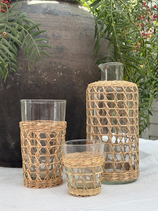 Three clear glass drinking vessels and a tall bottle, each wrapped in a natural woven rattan sleeve, displayed on a white tablecloth with a large dark planter and green foliage in the background.