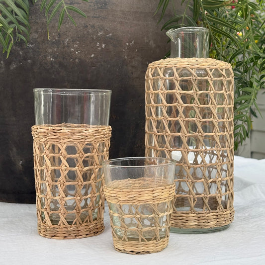 Three clear glass drinking vessels and a clear glass decanter with a woven natural rattan sleeve, arranged on a white tablecloth with a dark textured wall and green foliage in the background.