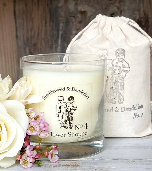 A white candle in a clear glass jar with greenery and flowers on the label, accompanied by a beige drawstring bag with a candle illustration, all displayed on a wooden surface.