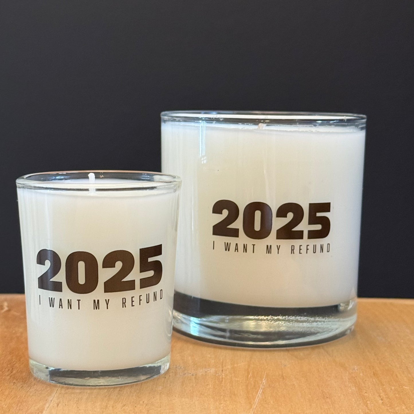 Two candles with '2025 I Want My Refund' text on a wooden surface.