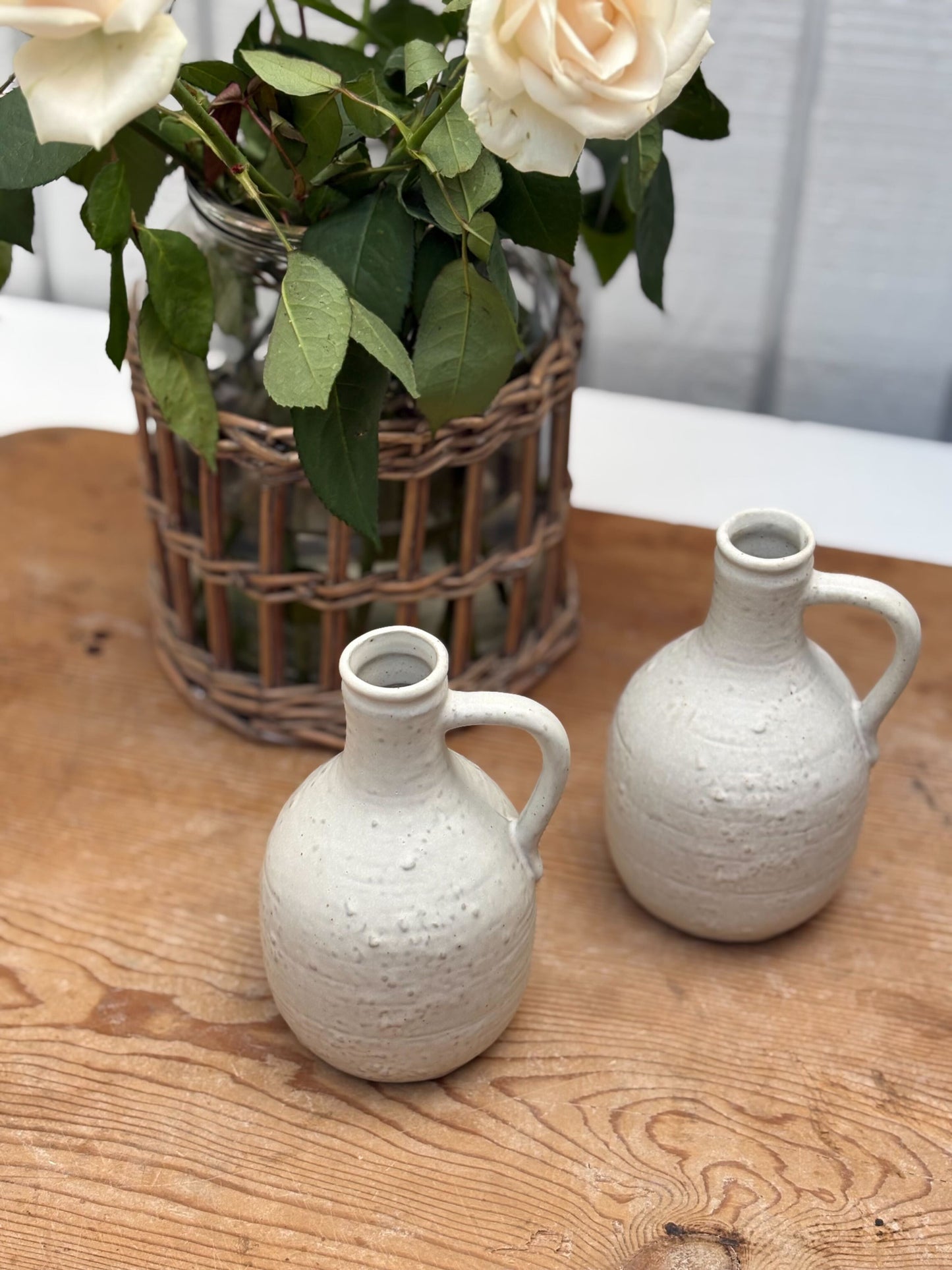 Two white ceramic jugs on a wooden table with a basket of flowers in the background.