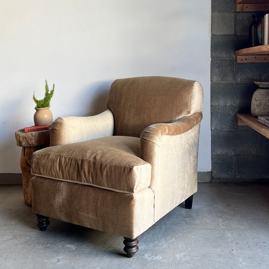 An upholstered chair with Suzani fabric back and taupe velvet body, featuring wooden legs, placed in a room with a plant and a shelf in the background.