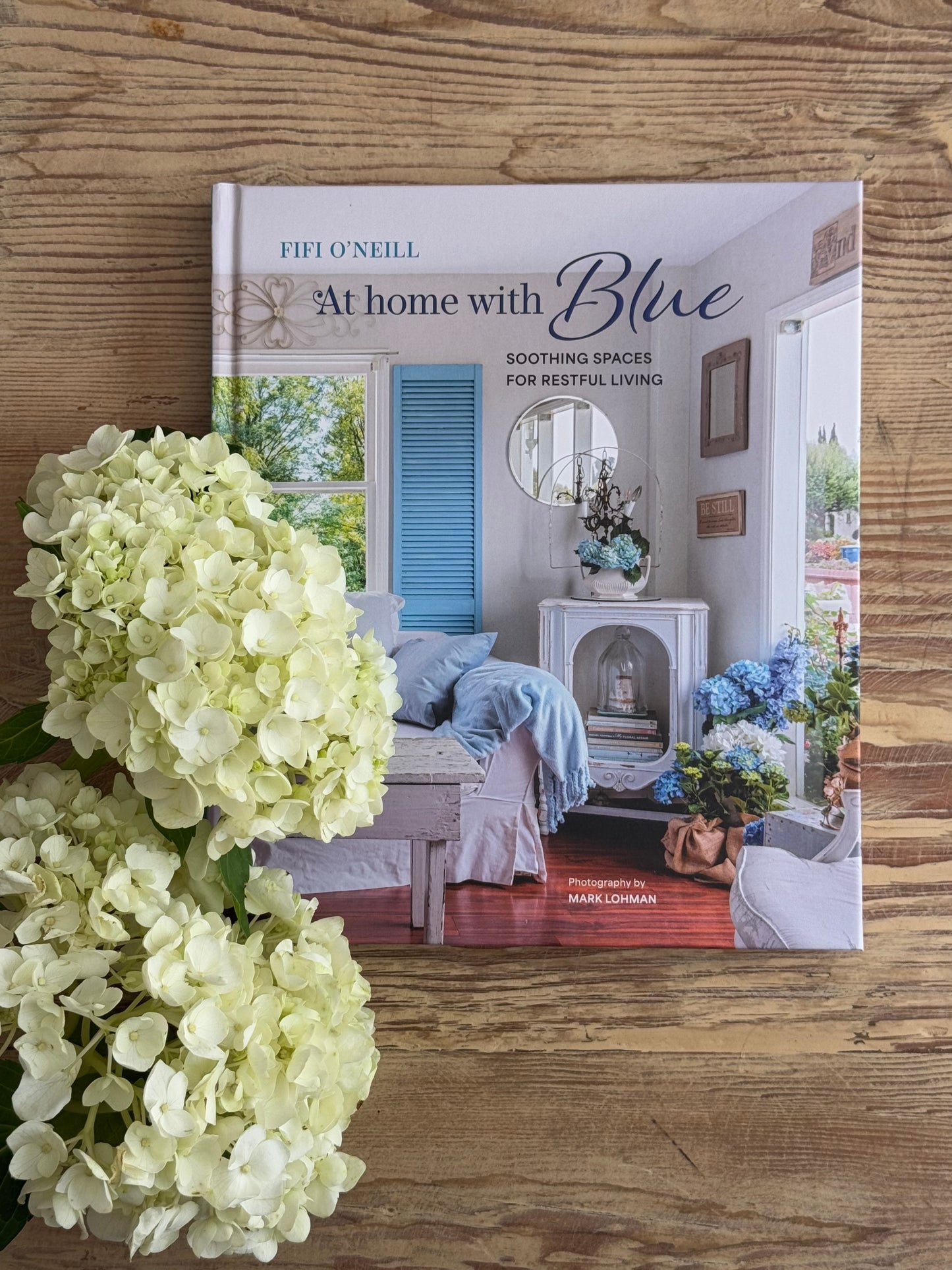 Book titled 'At Home with Blue' by Fifi O'Neill on a wooden surface with hydrangeas.
