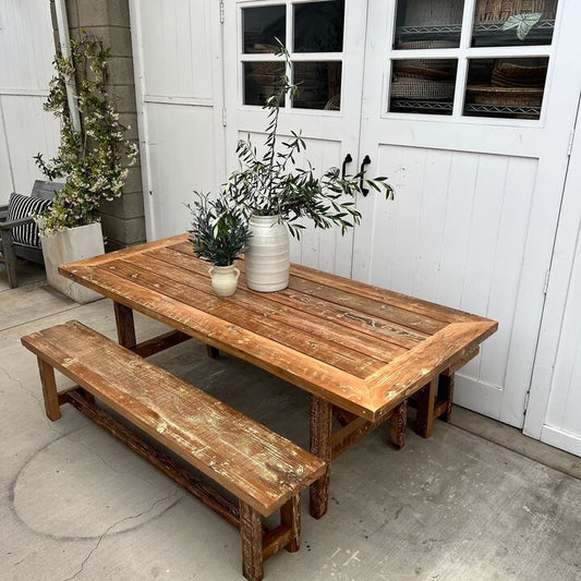 Wooden outdoor table with bench and potted plants against a white wall.