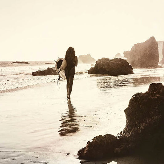 sepia toned photograph of woman with surfboard on beach with rocks