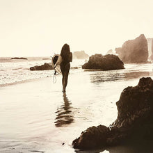 Load image into Gallery viewer, sepia toned photograph of woman with surfboard on beach with rocks