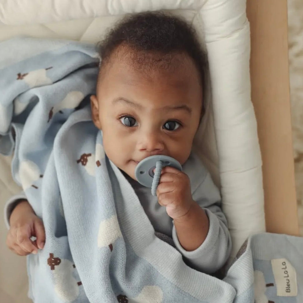 Baby lying in a crib with a pacifier, wrapped in a blue blanket with white patterns.