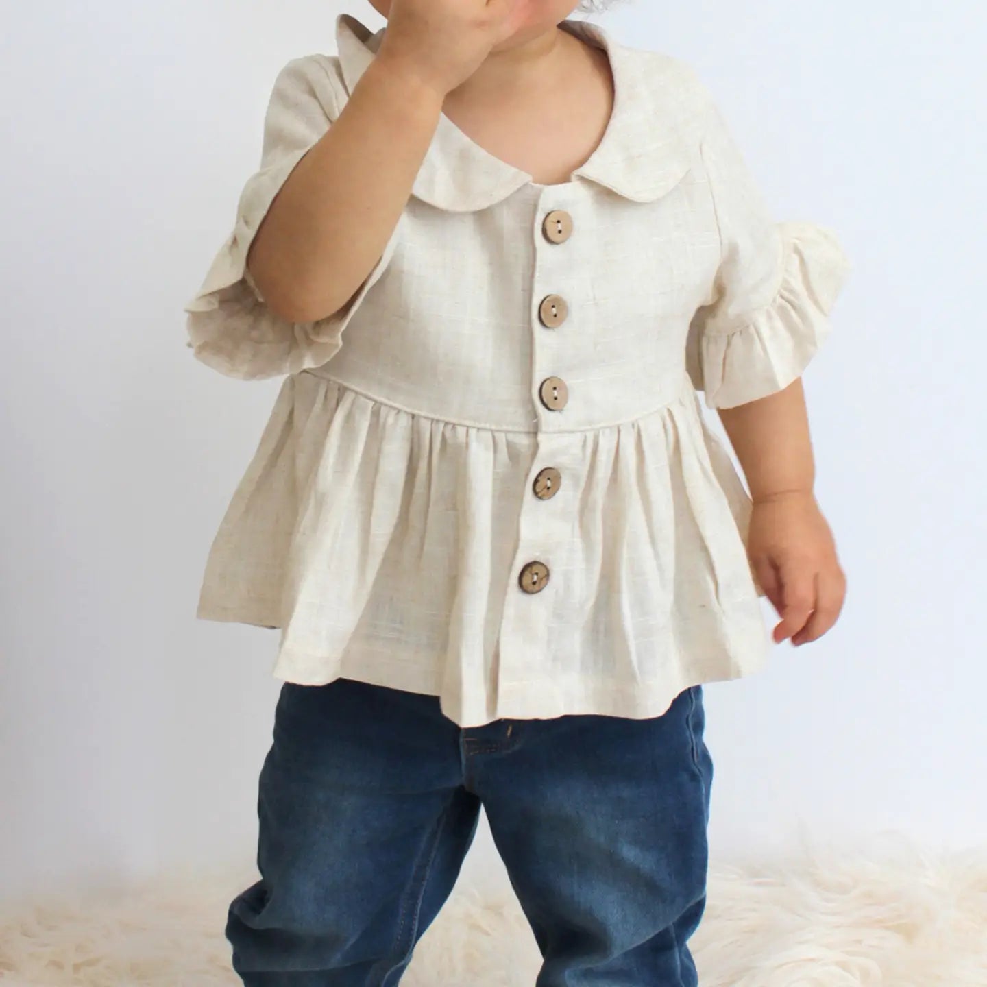 Child wearing a beige blouse with puffed sleeves and buttons, paired with blue pants on a light background.