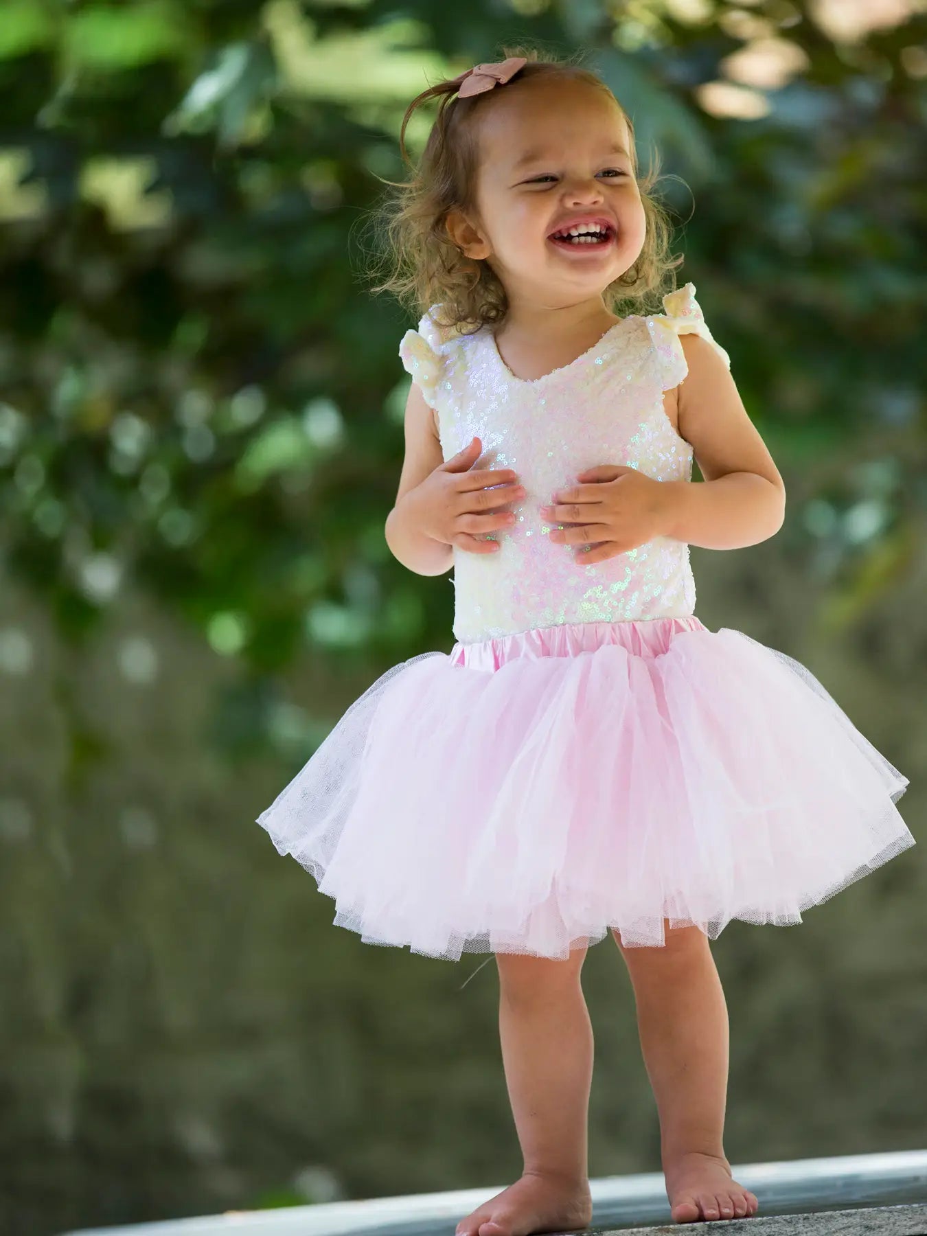 Child wearing a pink dress with a white lace top, standing outdoors with greenery in the background.