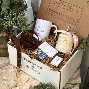 white gift box with: white mug that says "Best Dog Dad Ever", one glass Stinky Dog Candle, one brown leather dog leash, one package of sprinkled dog donuts, box of Tumbleweed matches