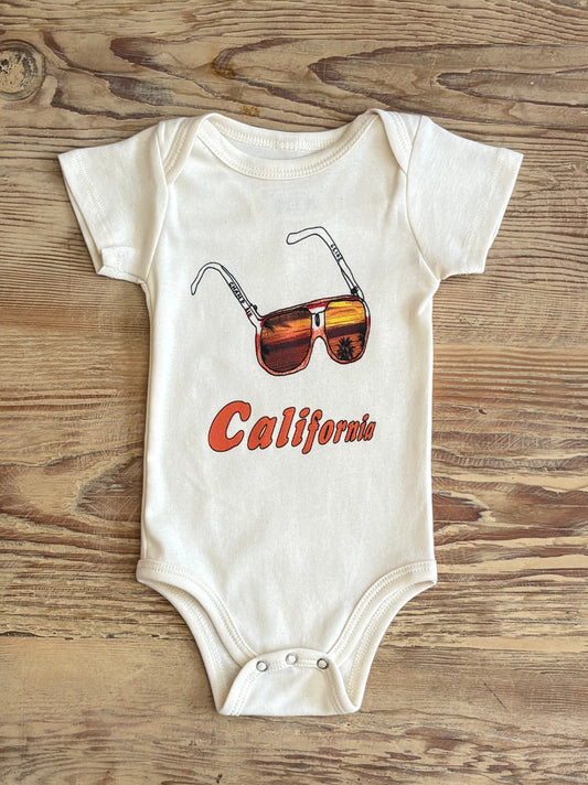 Baby onesie with sunglasses and 'California' text on a wooden surface