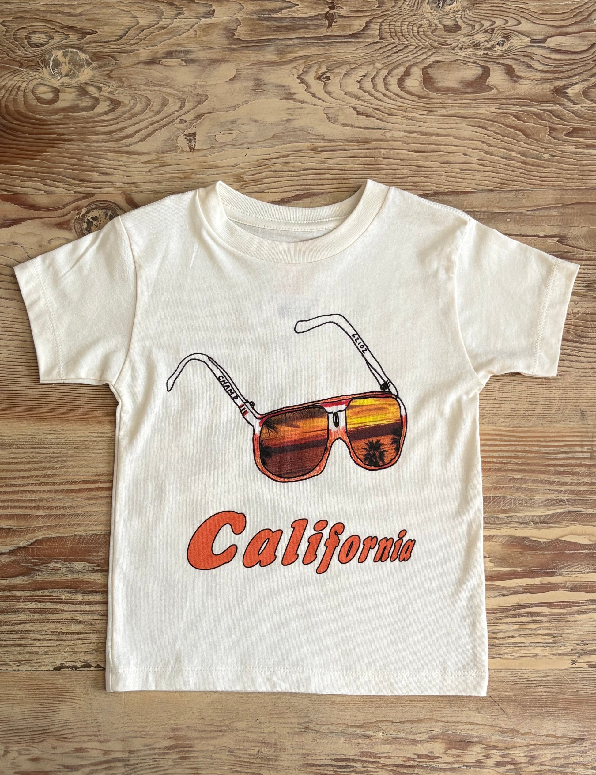 White t-shirt with sunglasses and 'California' text on a wooden surface