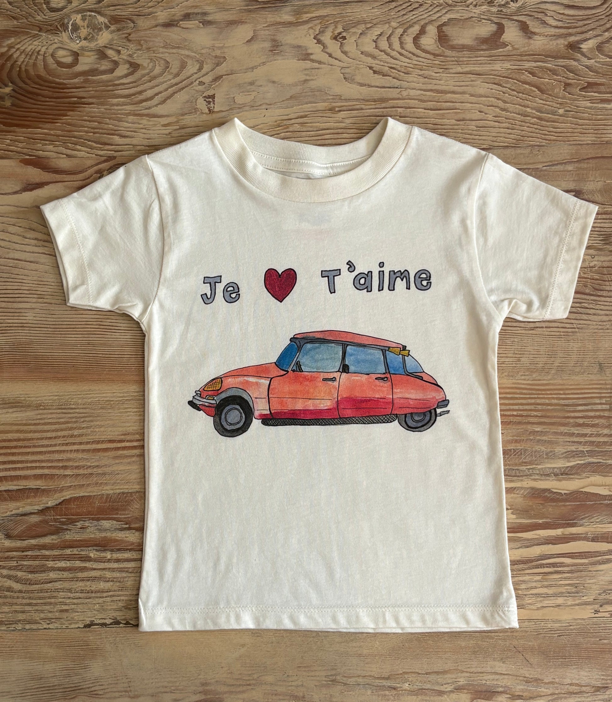 White t-shirt with a red car graphic and 'Je t'aime' text on a wooden surface