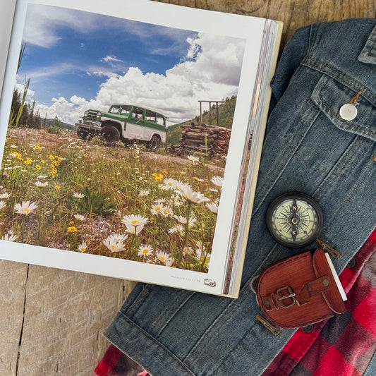 Open book with a photo of a green and white jeep in a field, next to a denim jacket and compass on a wooden surface.
