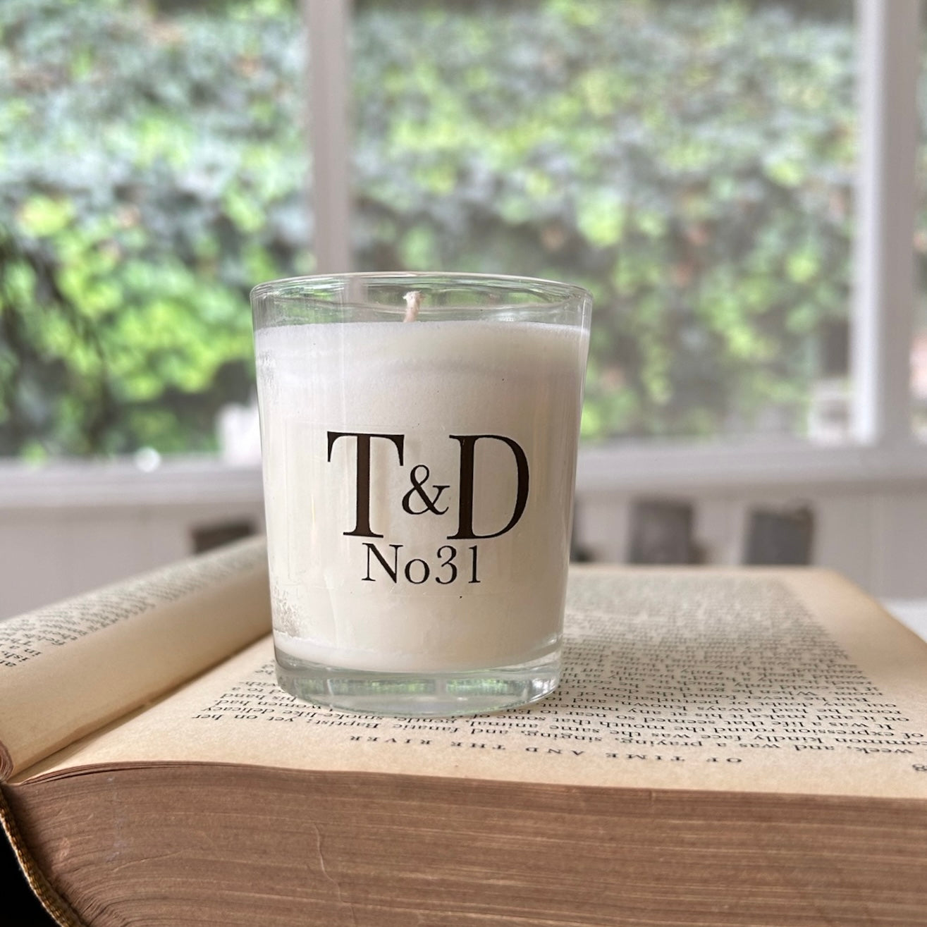 A white wax candle in a clear glass jar with black printed text reading 'T&D No31', placed on top of an open book in front of a softly blurred window with greenery outside.