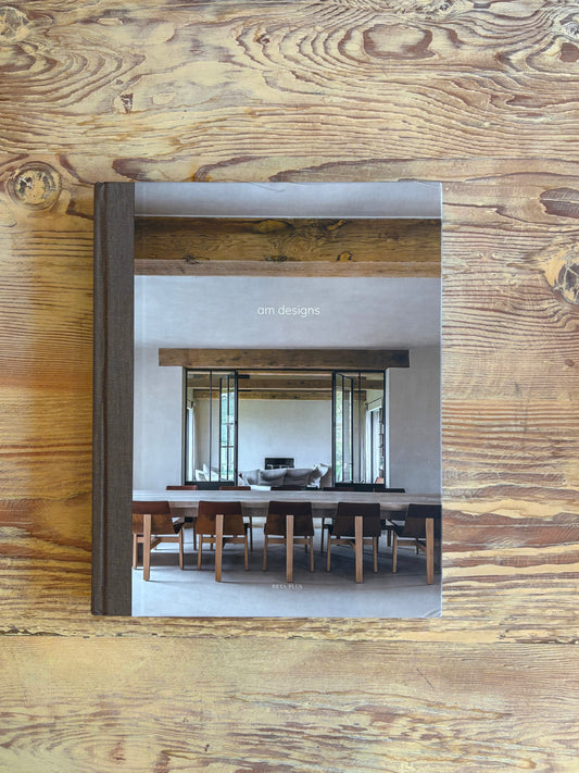 A hardcover book titled 'am designs' featuring a photograph of a modern dining room, displayed on a rustic wooden surface.