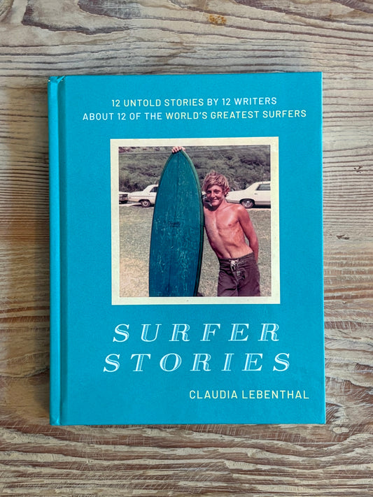 The book 'Surfer Stories' by Claudia Lebenthal, featuring a bright turquoise cover with white text and a central photograph of a shirtless surfer holding a blue surfboard, displayed on a rustic wooden surface.