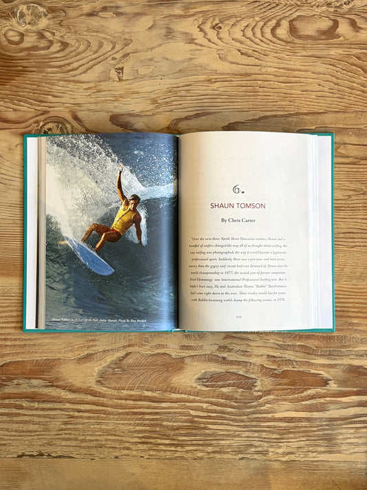 An open book laid flat on a rustic wooden table, showing a printed page with a photograph of a surfer on the left and text headed '6. SHAUN TOMSON' on the right.