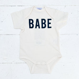 off white baby onesie with BABE in black on front