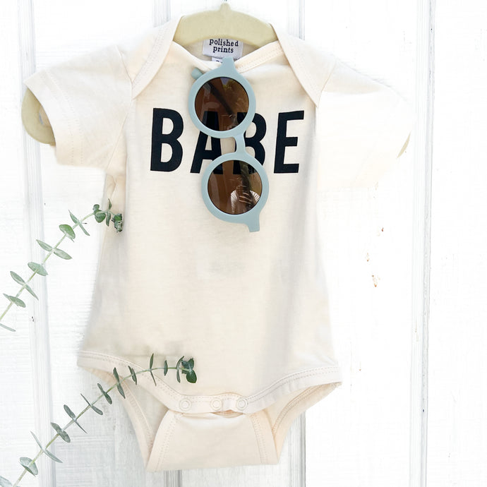 off white baby onesie with BABE in black on front