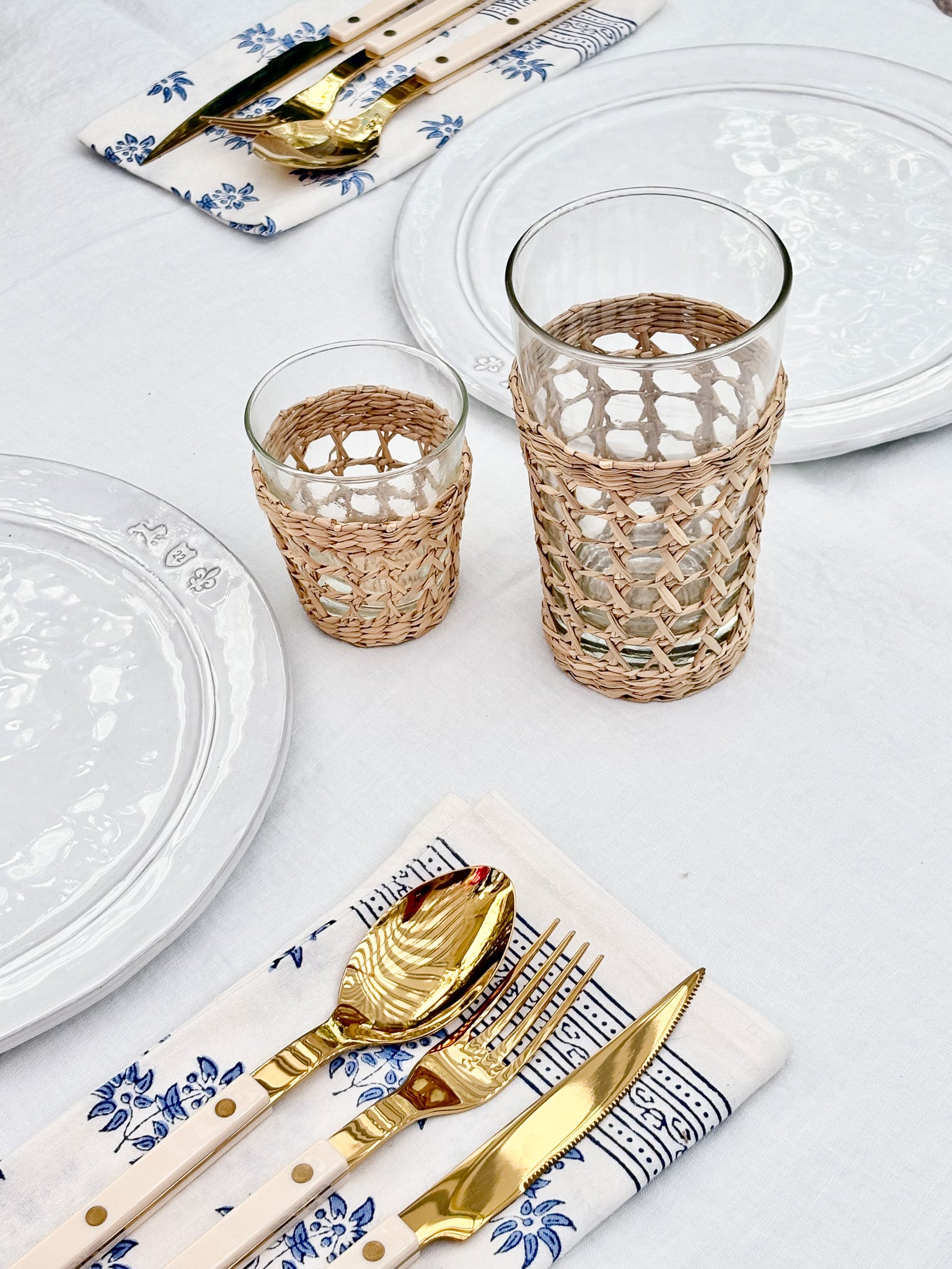 Two clear glass tumblers with woven natural-fiber sleeves, styled on a white table with white plates, gold flatware, and blue-and-white napkins.