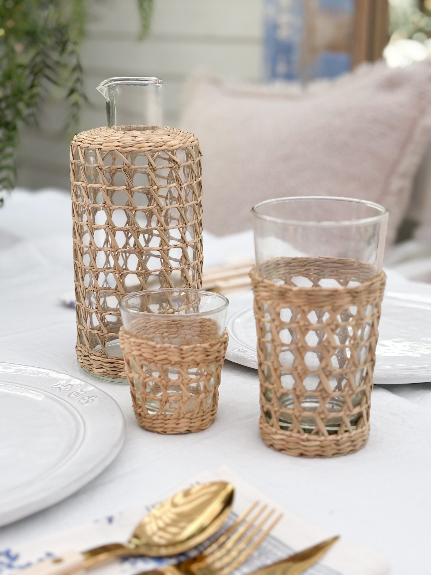 Three clear glass drinking vessels with woven natural-fiber sleeves in different heights, arranged on a white tablecloth with plates and gold-toned cutlery in a softly blurred indoor setting.