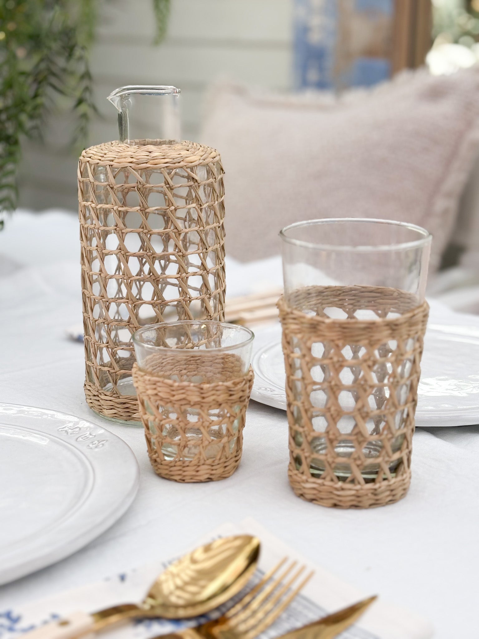 Three clear glass drinking vessels with woven natural-fiber sleeves in different heights, arranged on a white tablecloth with plates and gold-toned cutlery in a softly blurred indoor setting.