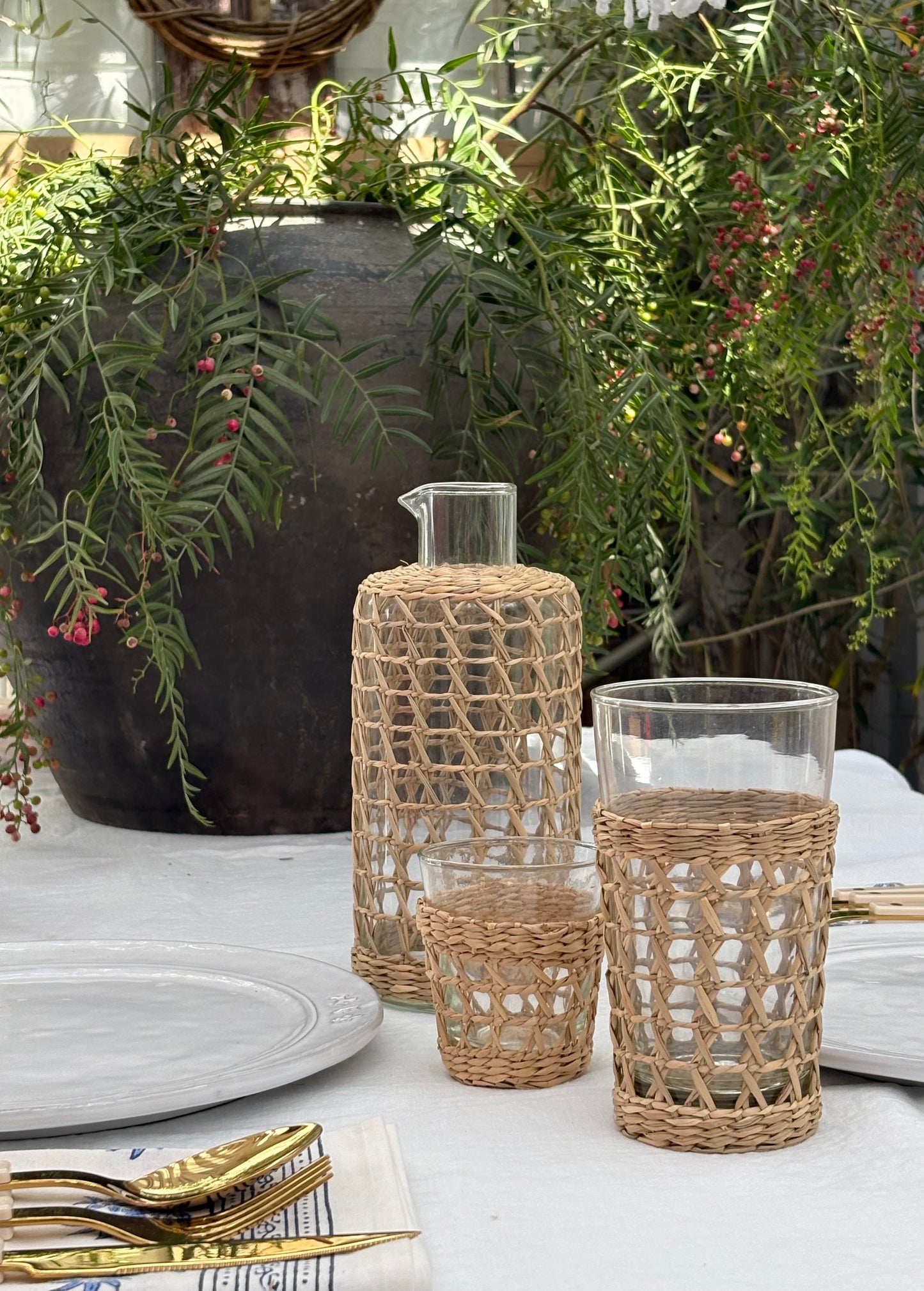 A set of clear glass drinking vessels with woven natural fiber covers, including a carafe and two tumblers, styled on a white tablecloth with gold cutlery and green foliage in the background.