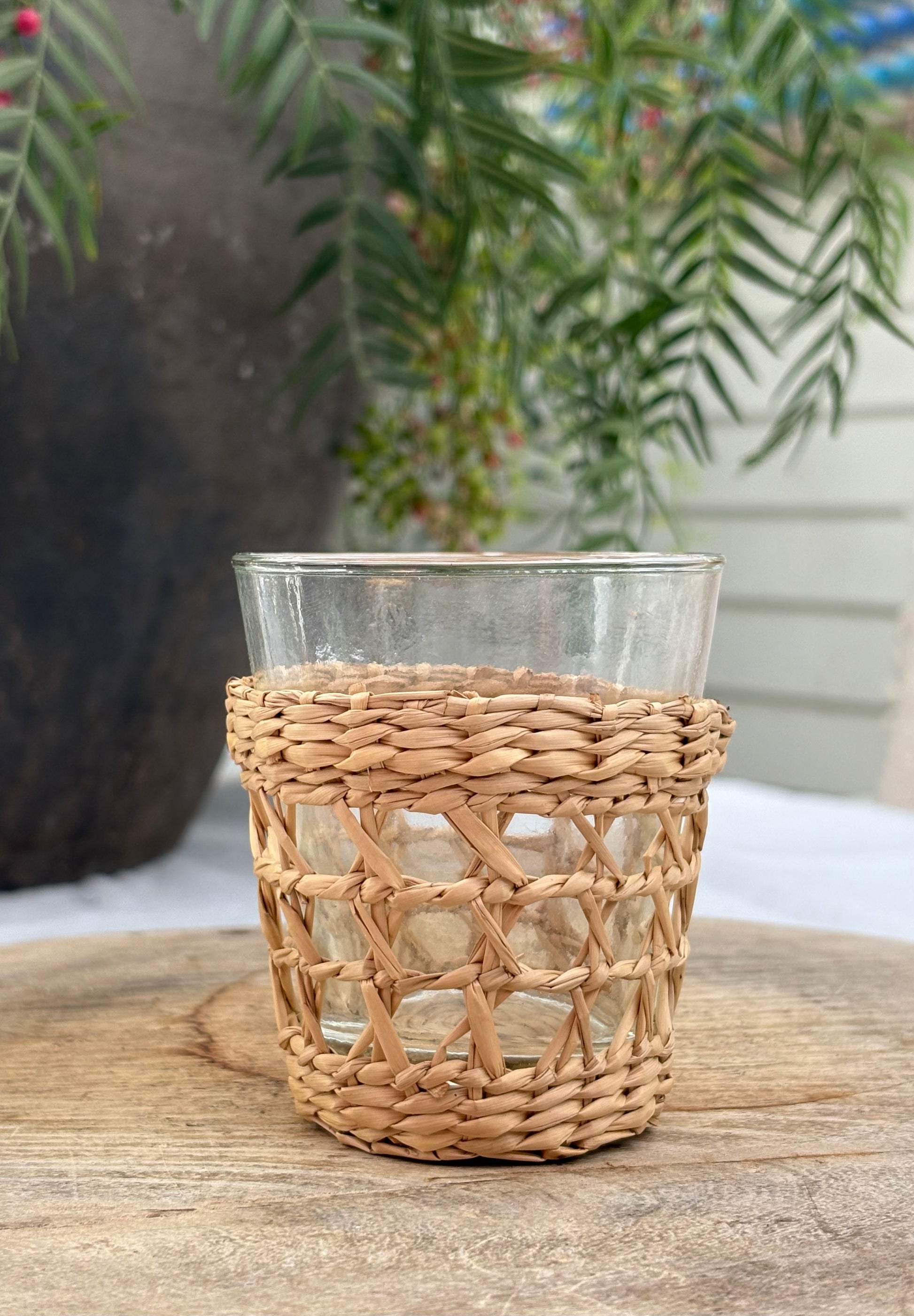 A clear glass tumbler with a natural woven straw sleeve, sitting on a rustic wooden surface with a blurred garden background.
