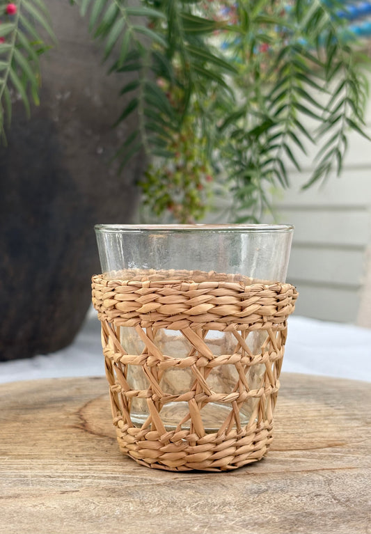 A clear glass tumbler with a natural woven straw sleeve, sitting on a rustic wooden surface with a blurred garden background.