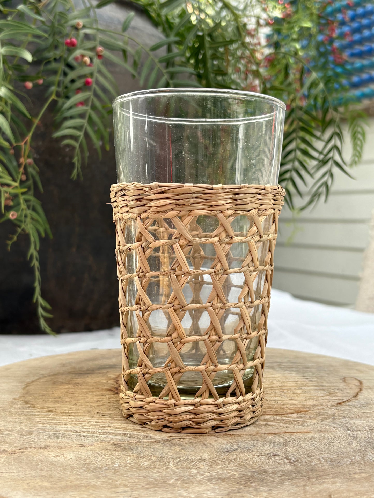 A clear glass tumbler with a natural woven rattan or wicker sleeve, displayed on a rustic wooden surface with green foliage in the background.