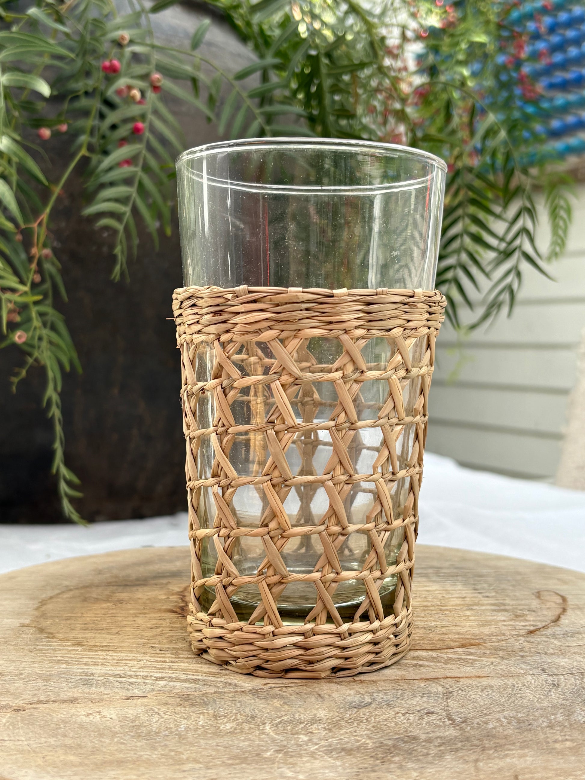 A clear glass tumbler with a natural woven rattan or wicker sleeve, displayed on a rustic wooden surface with green foliage in the background.