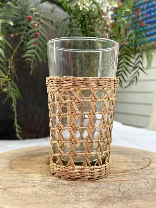 A clear glass tumbler with a natural woven rattan or wicker sleeve, displayed on a rustic wooden surface with green foliage in the background.