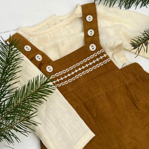 ochre colored corduroy baby overalls with cream buttons and cream stitching and long sleeved cream cotton shirt