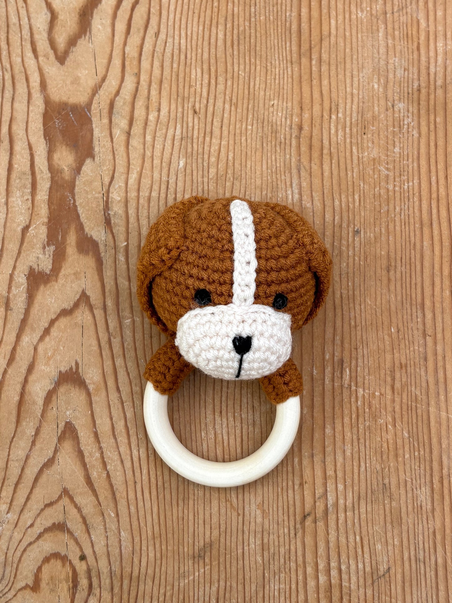 A crocheted dog-face baby teething ring with a brown and white dog design, attached to a cream-colored circular ring, displayed on a rustic wooden background.