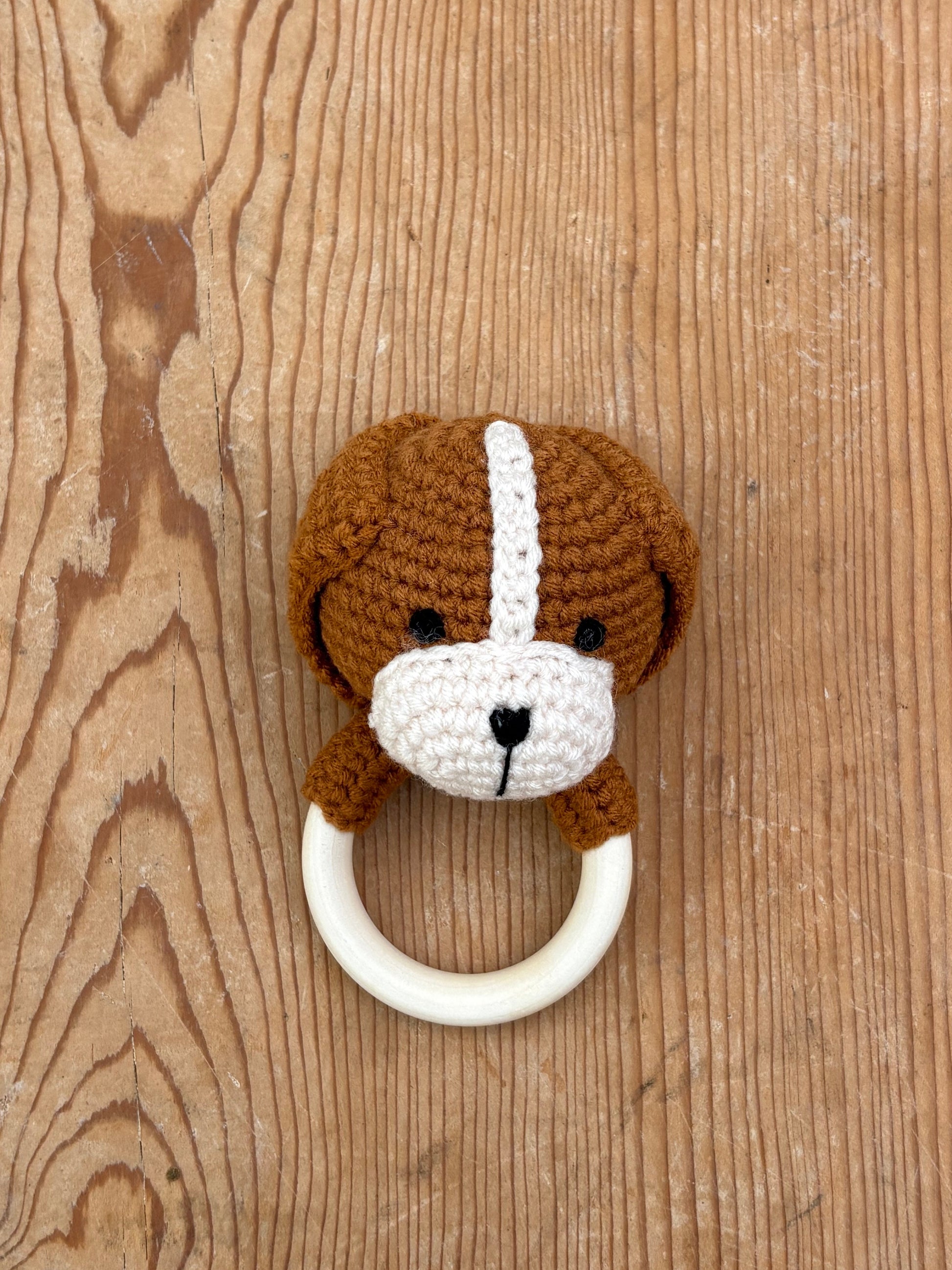 A crocheted dog-face baby teething ring with a brown and white dog design, attached to a cream-colored circular ring, displayed on a rustic wooden background.