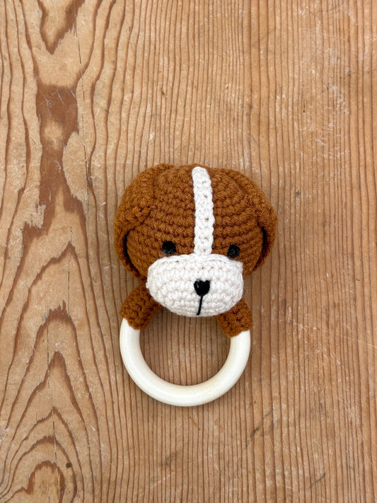 A crocheted dog-face baby teething ring with a brown and white dog design, attached to a cream-colored circular ring, displayed on a rustic wooden background.