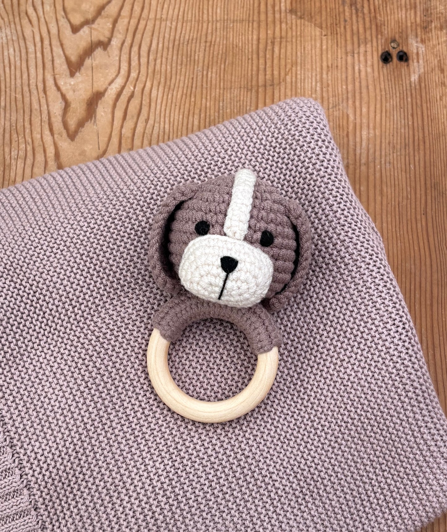 A crocheted dog-shaped baby teething ring toy with a wooden ring, resting on a knitted mauve blanket over a wooden surface.
