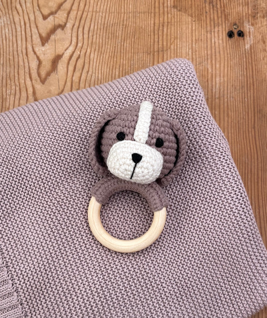 A crocheted dog-shaped baby teething ring toy with a wooden ring, resting on a knitted mauve blanket over a wooden surface.
