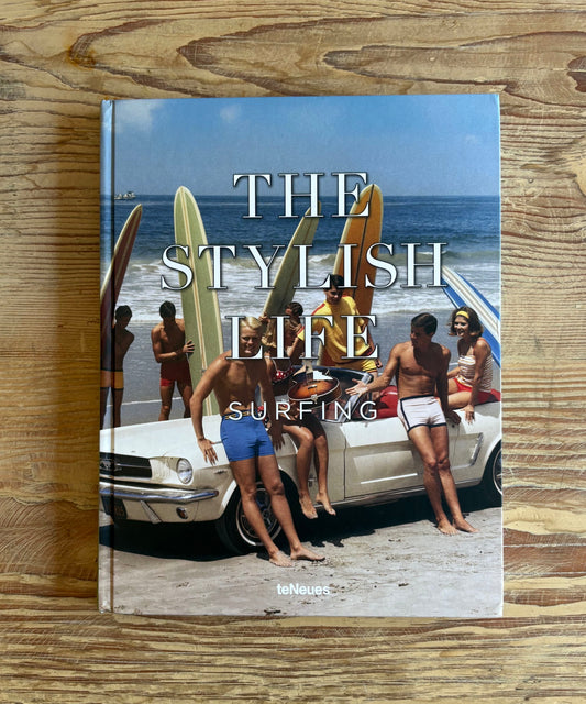 A hardcover book titled 'The Stylish Life of Surfing' by Melvyn Bragg, featuring a vintage beach photograph on the cover, displayed flat on a rustic wooden surface.