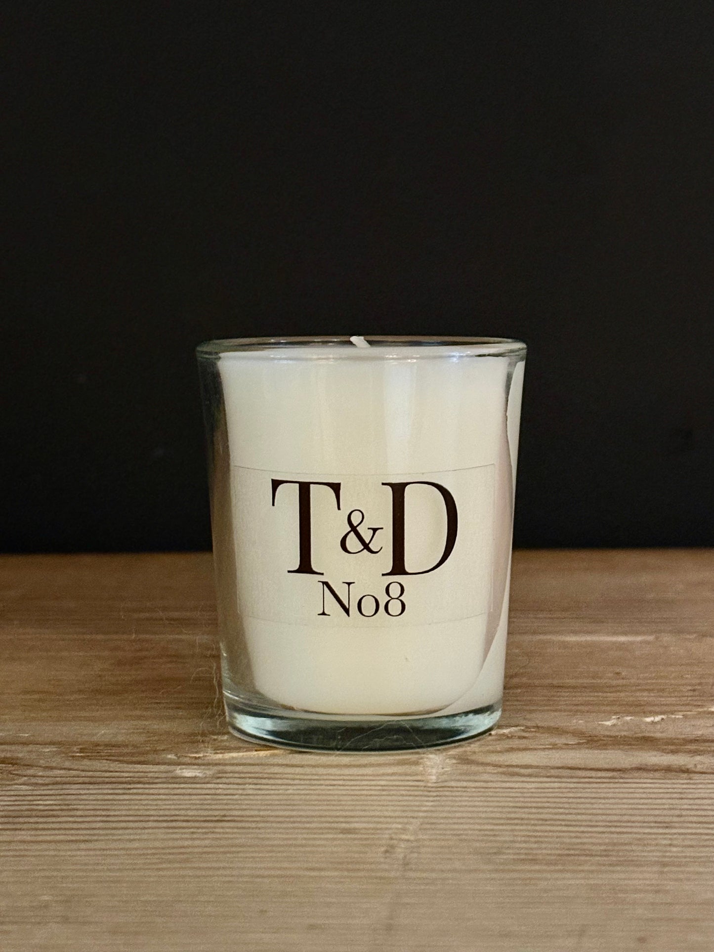 A white scented candle in a clear glass jar with black printed text reading 'T&D No8', placed on a light wood surface against a dark background.