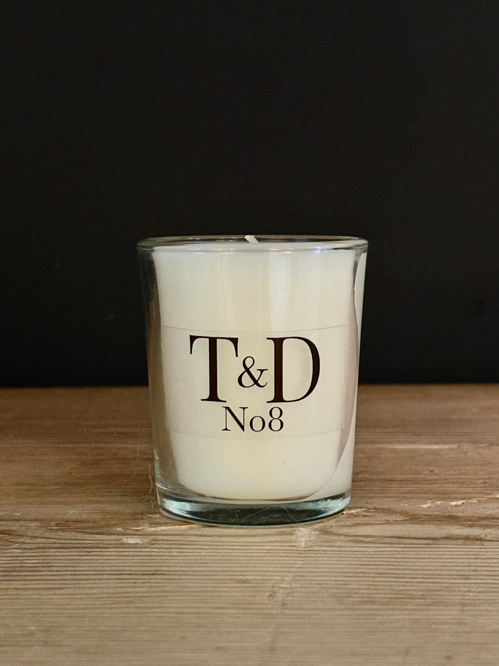 A white scented candle in a clear glass jar with black printed text reading 'T&D No8', placed on a light wood surface against a dark background.